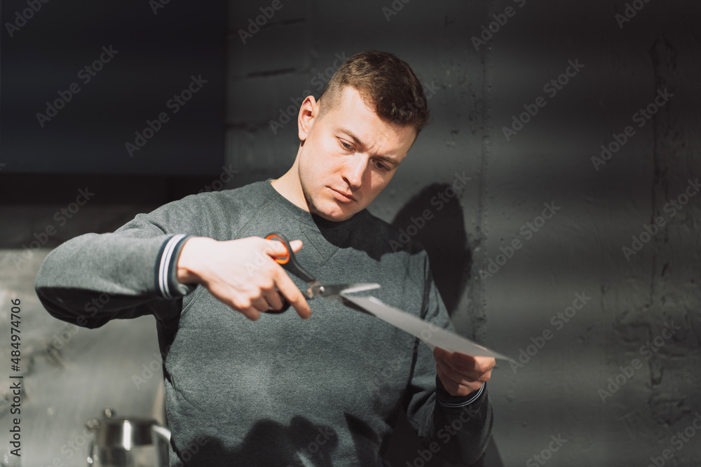a man cuts a white sheet of paper with scissors against a gray wall