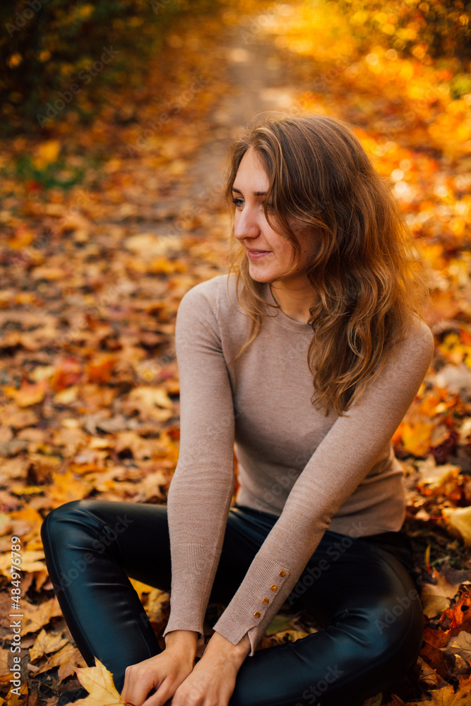 Beautiful girl is sitting on yellow leaves in the park. Attractive woman wearing beige sweater and black pants is walking in autumn park. Bright, warm sunny autumn day with yellow background.
