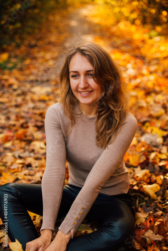 Beautiful girl is sitting on yellow leaves in the park. Attractive woman wearing beige sweater and black pants is walking in autumn park. Bright, warm sunny autumn day with yellow background. 
