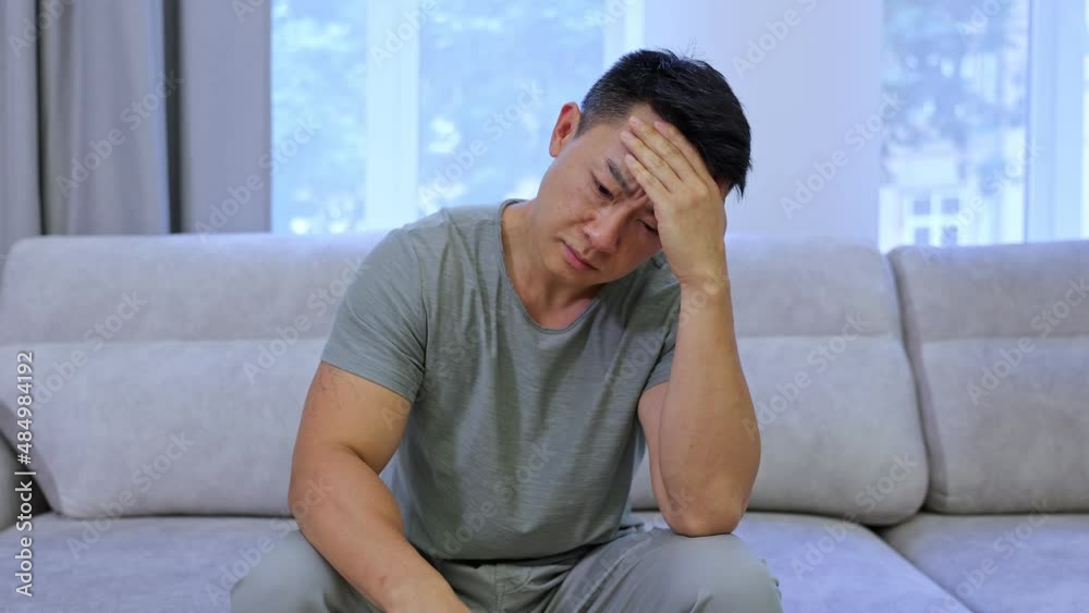 A tense man with a headache sits on the couch at home and rubs his temples. Upset man suffering from migraine or suffering from hangover touching his head A tense man with a headache sits on the couch at home and rubs his temples. Upset man suffering from migraine or suffering from hangover touching his head
