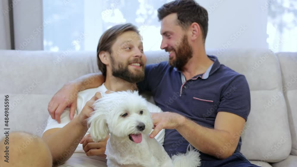 A happy gay couple is sitting on a couch at home and playing with a white dog.