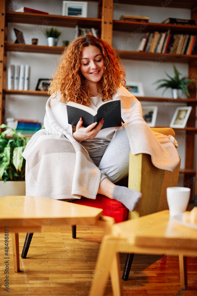 A redhead reading book at home. A happy ginger girl sits in an armchair covered with a blanket and reads an interesting book at her cozy home. A redhead reading book at home. A happy ginger girl sits in an armchair covered with a blanket and reads an interesting book at her cozy home.