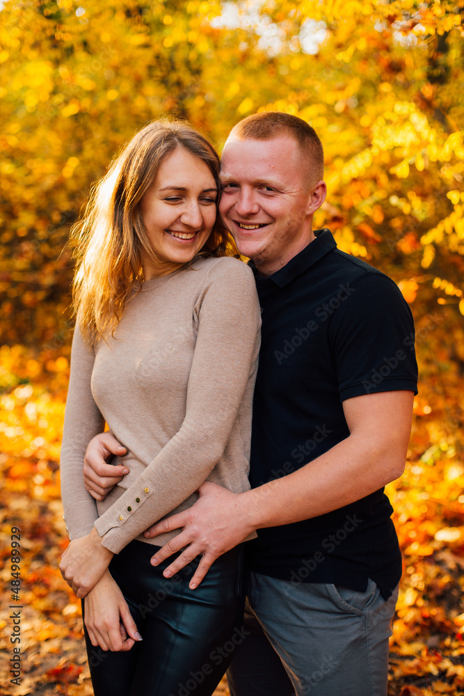 Fototapeta premium Young couple is having a walk in the sunny autumn park, forest. Happy man and his woman are hugging . Fallen yellow leaves, brith warm autumn weather. Autumn love story in park 