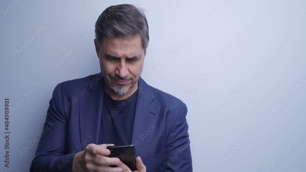 Business portrait - happy smiling businessman leaning to white wall using smart phone . Mature age, middle age, mid adult man in 50s with confident smile. Copy space. Business portrait - happy smiling businessman leaning to white wall using smart phone . Mature age, middle age, mid adult man in 50s with confident smile. Copy space.