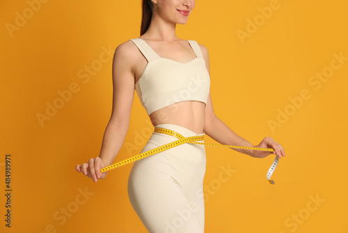 Happy woman in sportswear measuring waist with tape on yellow background, closeup