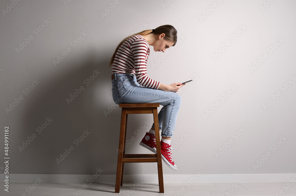 Woman with bad posture using tablet while sitting on stool near light grey wall indoors - obrazy, fototapety, plakaty 