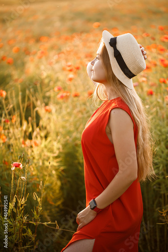 Happy cheerful woman walking on blooming field poppies. Red flowers. A blonde in a red short dress and a hat.