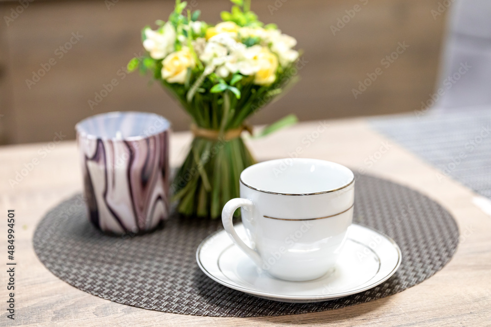 White mug and saucer set. Empty porcelain coffee cup next to decorative flowers and candlestick on wooden table in kitchen. Selective focus White mug and saucer set. Empty porcelain coffee cup next to decorative flowers and candlestick on wooden table in kitchen. Selective focus