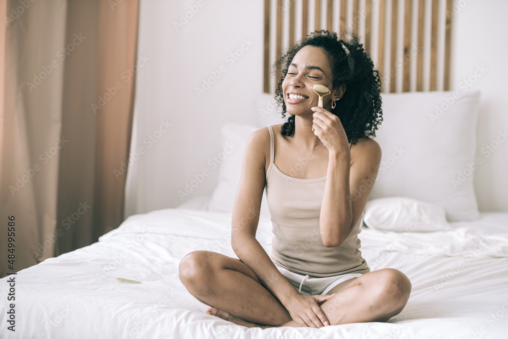 Morning routine concept. Happy young mixed race woman sitting on the bed using stone facial roller. Morning routine concept. Happy young mixed race woman sitting on the bed using stone facial roller.