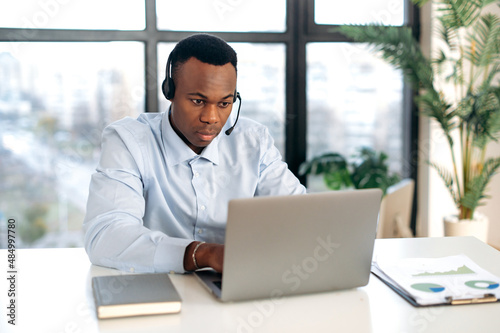 Smart purposeful, focused African American young man in headphones, manager, support worker, online consultant, sitting at a laptop in the office, having an online consultation with clients in a chat