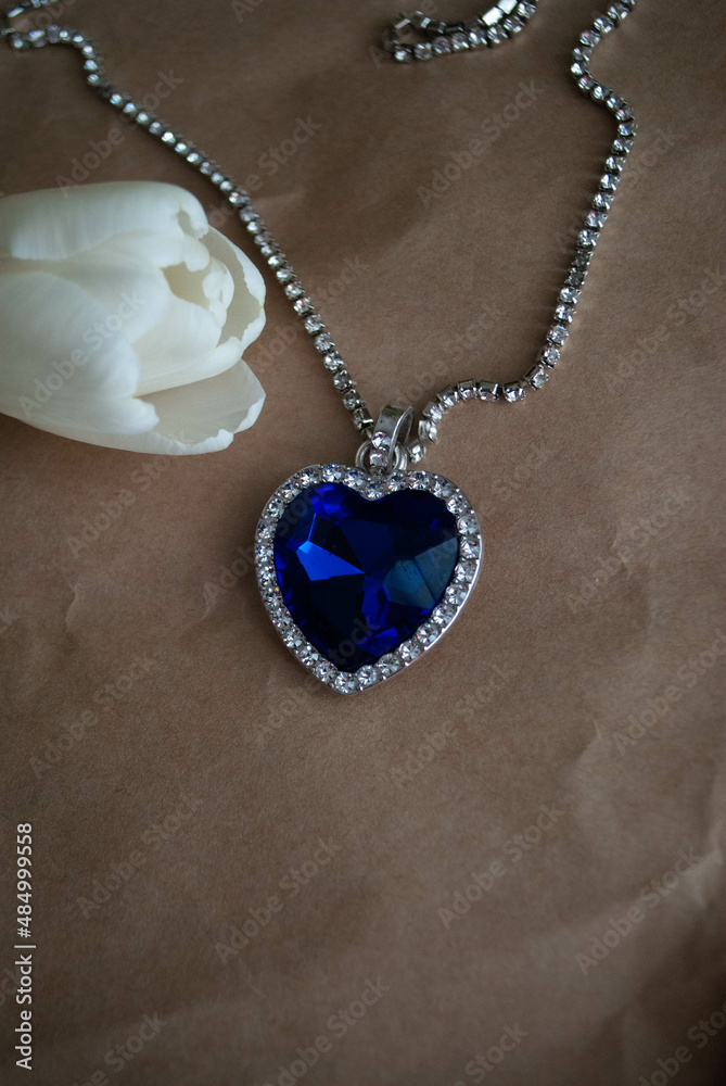 Close-up of the Heart of The Ocean necklace with white tulips bouquet on a craft paper. Titanic necklace. Blue diamond in a heart-shaped. Diamond necklace. Close-up of the Heart of The Ocean necklace with white tulips bouquet on a craft paper. Titanic necklace. Blue diamond in a heart-shaped. Diamond necklace.