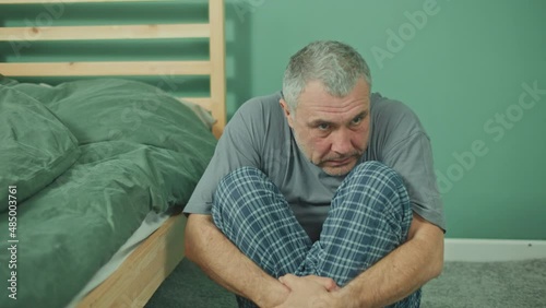 A Frightened Man Sitting on the Bedroom Floor Next to the Bed, Suffering from Depression and Paranoia, Feeling Overwhelmed and Helpless. Panic Attack. Medical Services and Mental Health Concept. photo