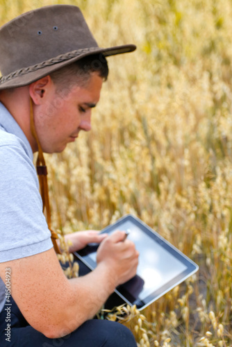 Digital farm. Portrait of farmer seating in gold wheat field and scrolling on tablet. Young man wearing cowboy hat in field examining wheat crop. Oats grain industry. Blurred. Oats plant. Copy space