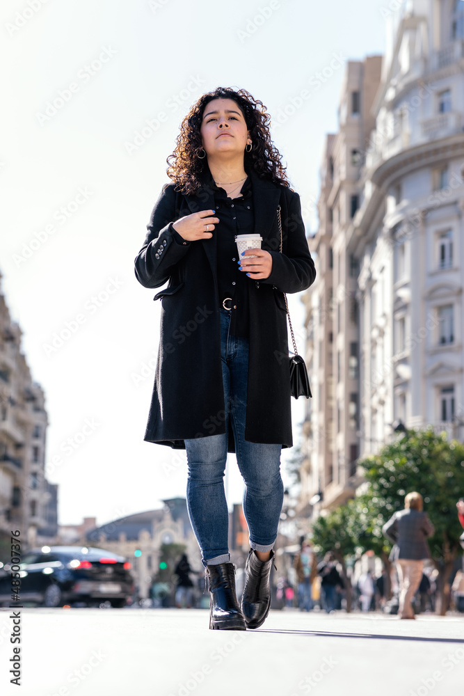 Fototapeta premium Beautiful and young elegant woman walking with coffee in hand through the city on a sunny day