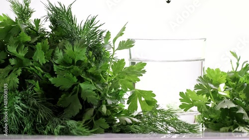Chlorophyll extract is poured in pure water in glass against a white background and green organic dill and parsley herbs. Growing fresh plants, healthy food. Concept of detox diet, slow motion photo