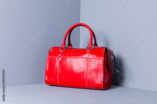 A bright red tote bag stands on a wooden gray background. The bag is decorated with white stitching