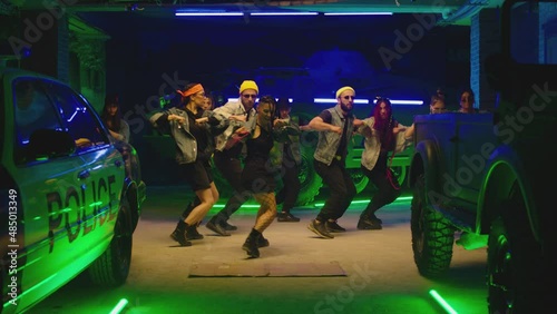 Group of stylish young people dancing synchronized dance  , moving near military tank and police car inside garage building Colorful stylish clothes  Modern professional dances . Green blue led lights