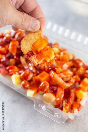 A view of a hand holding a chip full of toppings from a plastic container of tostilocos. photo