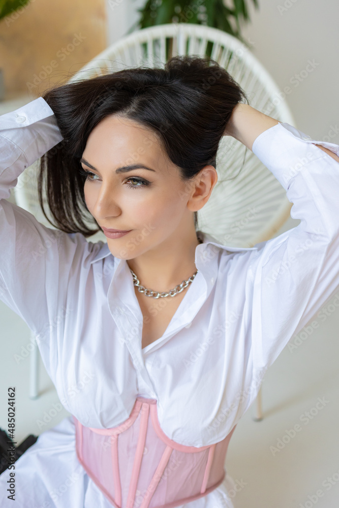 brunette girl in a white shirt-dress and a pink corsage sits in a wicker chair brunette girl in a white shirt-dress and a pink corsage sits in a wicker chair