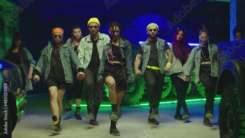 Group of stylish young people dancing synchronized dance  , moving near military tank and police car inside garage building Colorful stylish clothes  Modern professional dances . Green blue led lights