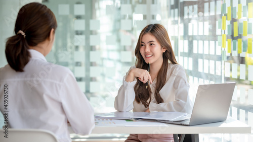 Two pretty young Asian businesswoman sitting at desk with laptop doing paperwork together discussing project financial report. Corporate business collaboration concept.