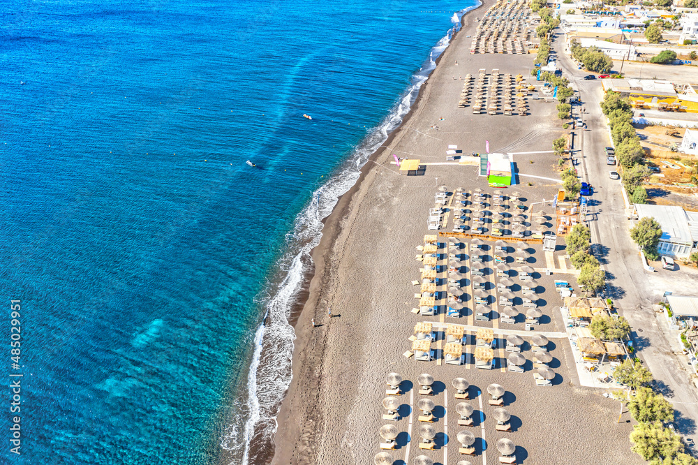 Top view aerial drone photo of black Perissa beach with beautiful turquoise water, sea waves and straw umbrellas. Vacation travel background. Aegean sea, Santorini Island, Greece