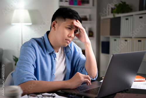Unhappy young Asian man, entrepreneur, student, tutor looking at laptop screen with serious and worried face and with hand on forehead when working or learning from home.