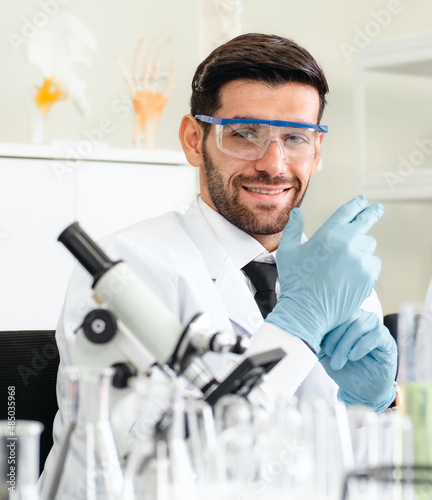 Male scientist is working in pharmaceutical and medical research lab with microscope and liquid in test tubes. Smiling doctor analyzing innovative virus protective vaccines in health care laboratory.