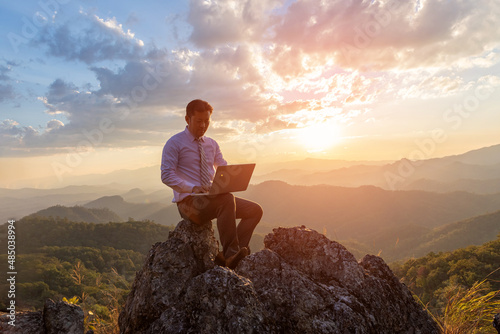 Business man working outdoors with laptop computer on top rock mountain at sunset background. Work and vacation.