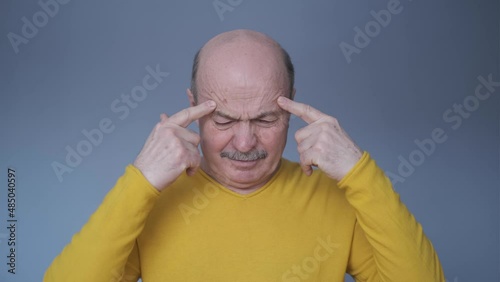Thoughtful senior man holding fingers on temples standing isolated on blue wall