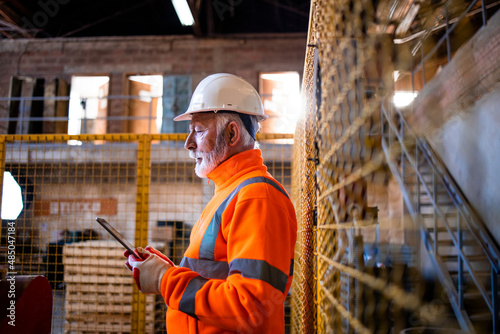 Factory worker or industrial supervisor in safety uniform and hard hat standing in factory production hall and using tablet computer.