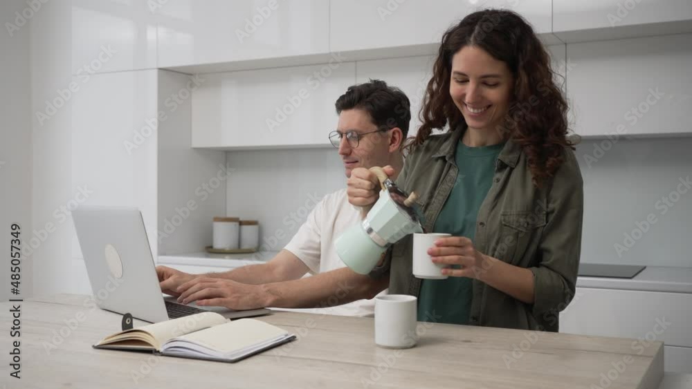 Man with headphones on neck frowns working distantly via laptop and brunette wife pours coffee into cups to cheer up husband smiling and kissing Man with headphones on neck frowns working distantly via laptop and brunette wife pours coffee into cups to cheer up husband smiling and kissing