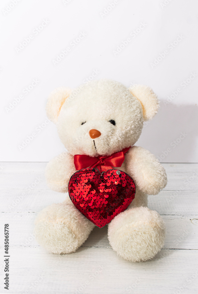 Image of white toy teddy bear sitting on white wooden background with red bow andl heart
