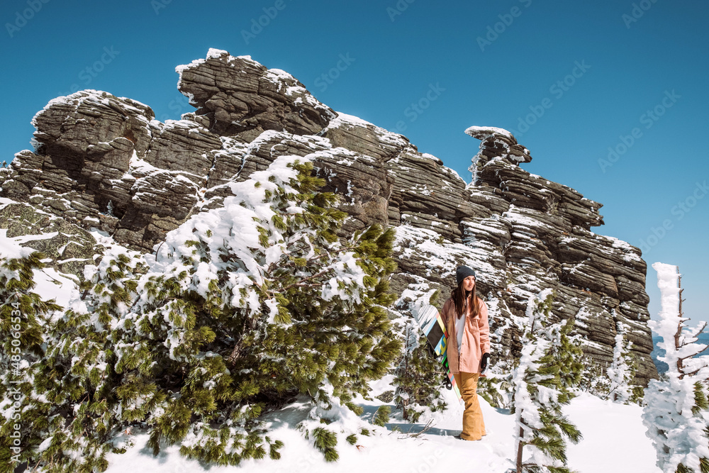 Russia. Sheregesh. Girl snowboarder in winter in sunny weather outdoors among the Christmas tree, huge rock and snow. The girl is standing with a snowboard in her hands and posing. 