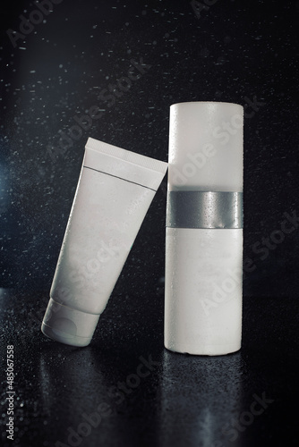 White bottles of cosmetics on a black background in water drops