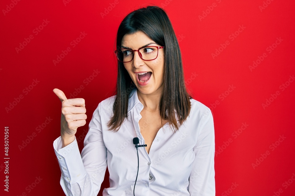 Young hispanic woman using lavalier microphone pointing thumb up to the side smiling happy with open mouth Young hispanic woman using lavalier microphone pointing thumb up to the side smiling happy with open mouth