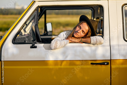 Caucasian Melancholic Or Sleepy Woman Is Leaned On Van Window Having Rest At Summer Evening, In yellow Camping Retro Van. Amazing Beautiful Female In Hat Is Relaxing. Trip, Travel Concept