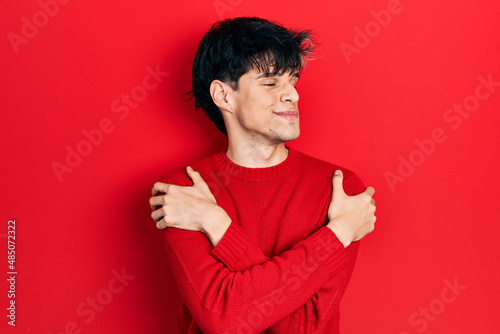 Handsome hipster young man wearing red winter sweater hugging oneself happy and positive, smiling confident. self love and self care © Krakenimages.com