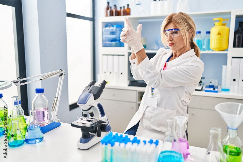 Middle age blonde woman working at scientist laboratory holding symbolic gun with hand gesture, playing killing shooting weapons, angry face Middle age blonde woman working at scientist laboratory holding symbolic gun with hand gesture, playing killing shooting weapons, angry face