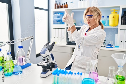Middle age blonde woman working at scientist laboratory holding symbolic gun with hand gesture  playing killing shooting weapons  angry face