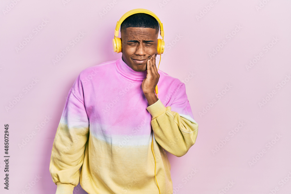 Fototapeta premium Young black man listening to music wearing headphones touching mouth with hand with painful expression because of toothache or dental illness on teeth. dentist