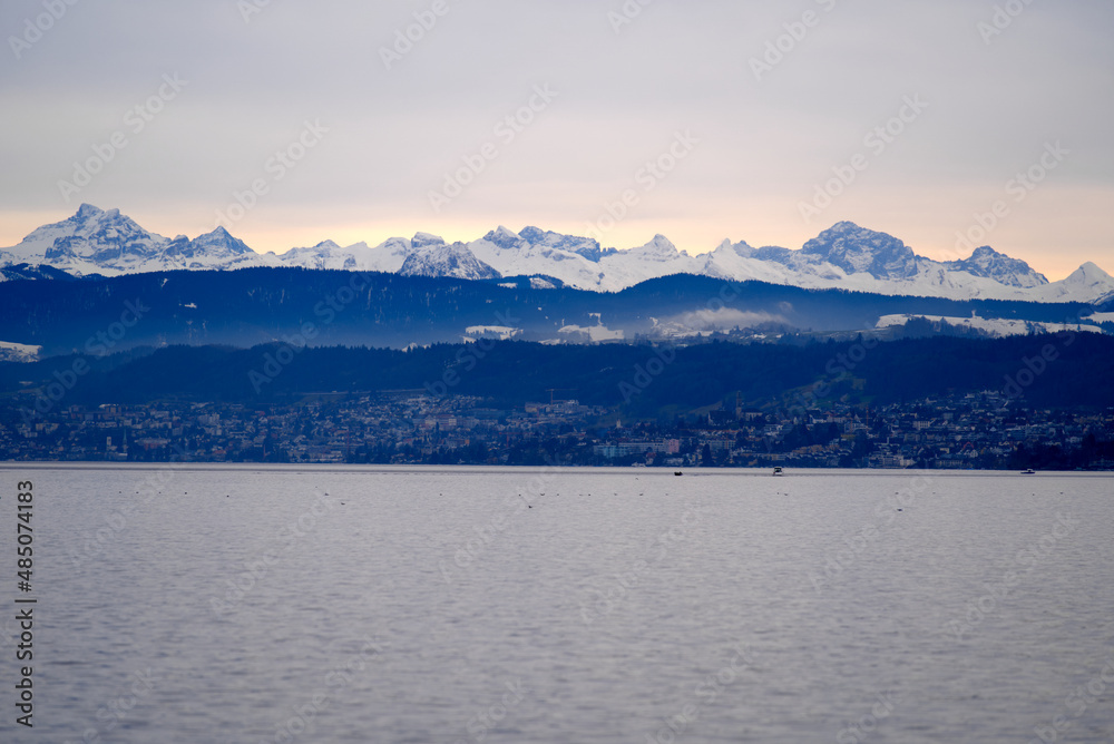 Beautiful scenic landscape with Lake Zurich in the foreground and Swiss Alps in the background on a cloudy winter afternoon. Photo taken February 3rd, 2022, Zurich, Switzerland.