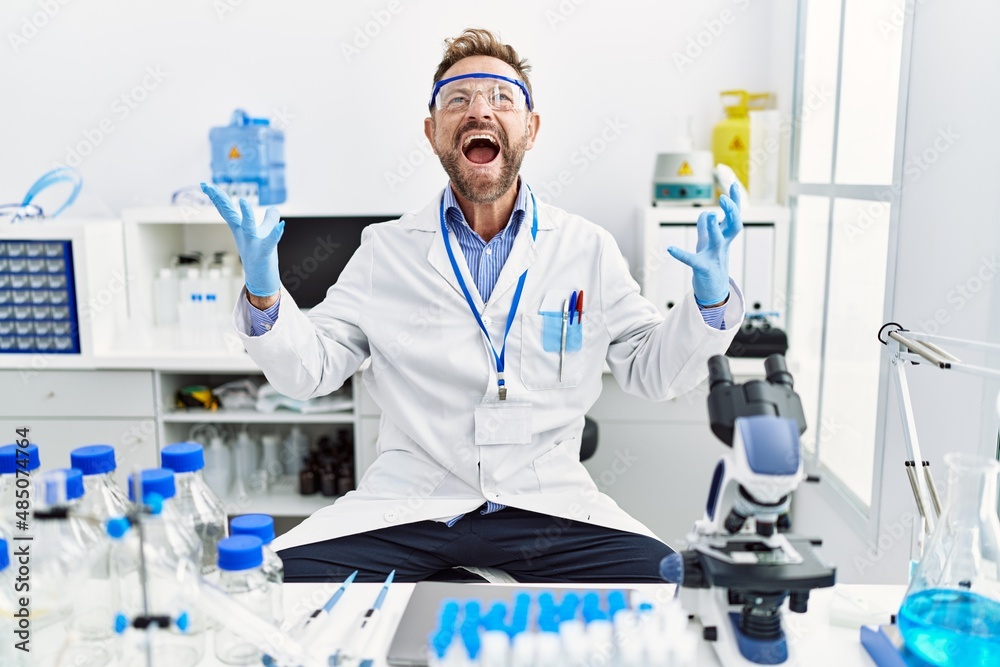 Middle age man working at scientist laboratory crazy and mad shouting and yelling with aggressive expression and arms raised. frustration concept.
