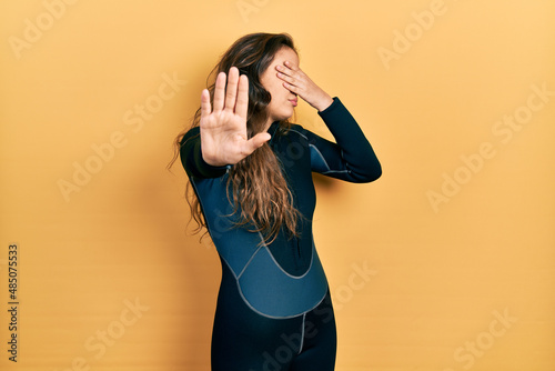 Young hispanic girl wearing diver neoprene uniform covering eyes with hands and doing stop gesture with sad and fear expression. embarrassed and negative concept.