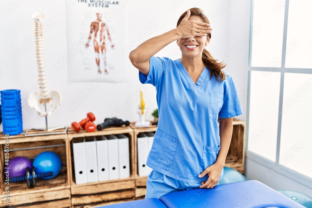 Middle age hispanic physiotherapist woman working at pain recovery clinic smiling and laughing with hand on face covering eyes for surprise. blind concept.