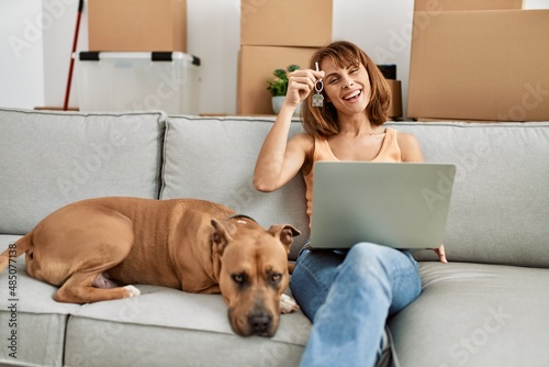 Young caucasian woman having video call and holding key sitting on sofa with dog at home