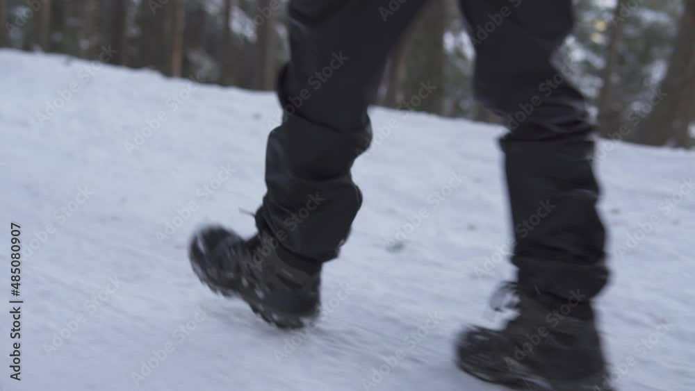 Shot showing man grey hiking boots while he walks outdoors on the snow and ice in the middle of the mountains, with a pine forest in the background. Diagonal side view Shot showing man grey hiking boots while he walks outdoors on the snow and ice in the middle of the mountains, with a pine forest in the background. Diagonal side view