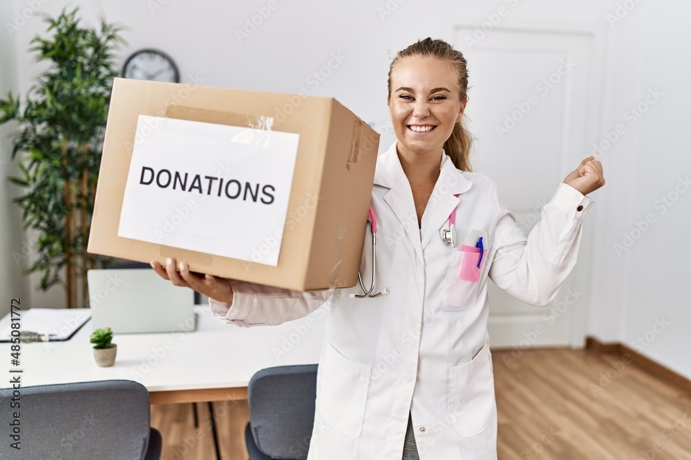 Young doctor woman at the clinic holding donations box screaming proud, celebrating victory and success very excited with raised arm Young doctor woman at the clinic holding donations box screaming proud, celebrating victory and success very excited with raised arm