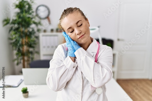 Young caucasian woman wearing doctor uniform and stethoscope at the clinic sleeping tired dreaming and posing with hands together while smiling with closed eyes.