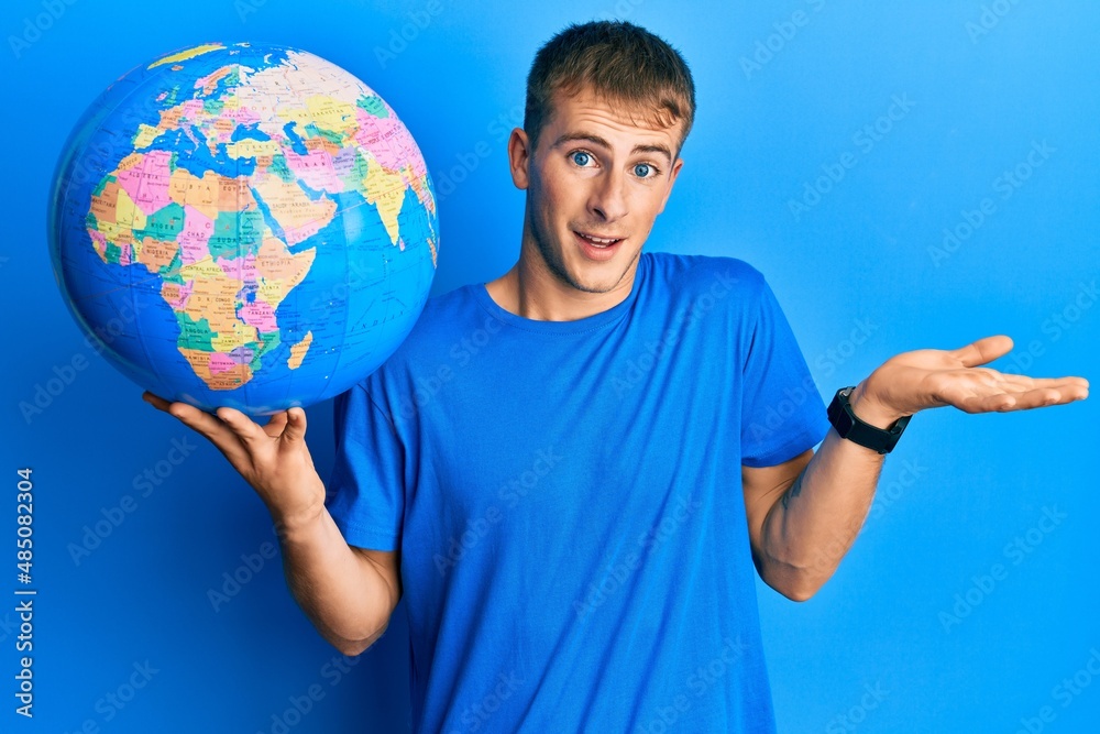Young caucasian man holding world ball celebrating achievement with happy smile and winner expression with raised hand Young caucasian man holding world ball celebrating achievement with happy smile and winner expression with raised hand